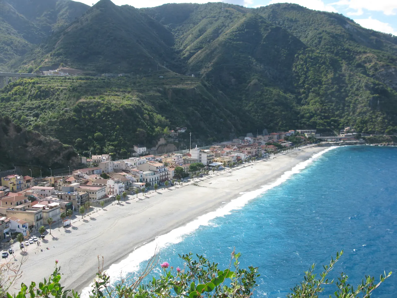 Marina Grande a Scilla, con il mare azzurro, le case lungo la spiaggia e i rilievi verdi alle spalle.